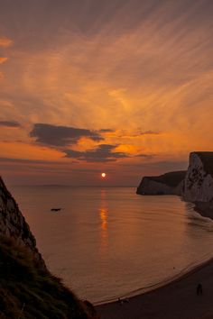 the sun is setting over the ocean with cliffs in the distance and people walking on the beach