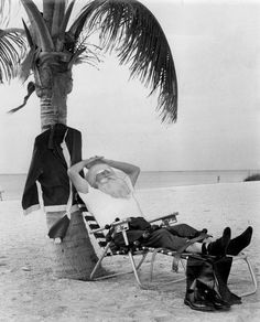 a man laying on top of a beach next to a chair under a palm tree