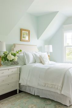 a white bed sitting under a window next to a dresser with flowers on top of it