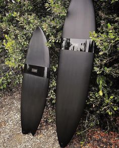 two black surfboards sitting next to each other in front of some bushes and shrubbery