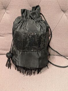 This is a stylish black drawstring purse with a vintage flair. The main body is made of a smooth, satin-like material, while the bottom features a band of shimmering sequins that add a touch of glamour. The purse has a drawstring closure and a tassel detail. The fringe trim at the bottom adds a bohemian touch. Sequin Handbag, Drawstring Purse, Simi Valley, Top Handle Bags, Fringe Trim, Purses And Handbags, Top Handle, Top Handle Bag, Bathing Beauties