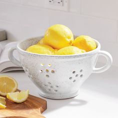 a white colander filled with lemons on top of a counter next to a cutting board