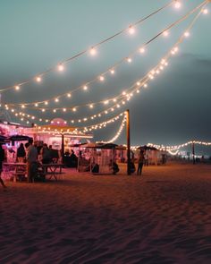 people are standing on the beach at night with lights strung over them and food stalls in the background