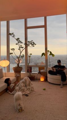 two people and a dog in a living room with large windows looking out on the city