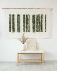 a wall hanging with plants on it in front of a white bench and wooden table