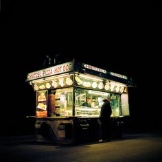 a man standing in front of a hot dog stand at night with the lights on