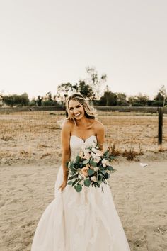 a woman standing in the sand with a bouquet on her shoulder and wearing a strapless wedding dress