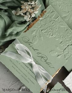 a green wedding card with a white ribbon and flower on the front is laying next to it's matching envelope