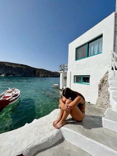 a woman sitting on the edge of a cliff next to some water with boats in the background