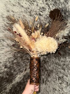 a person holding a vase with dried flowers and feathers in it on a furry surface