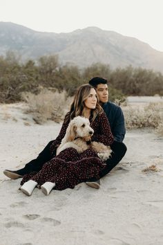 a man and woman sitting in the sand with a dog