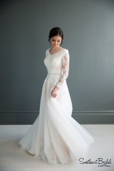 a woman in a white wedding dress standing against a gray wall with her hands on her hips