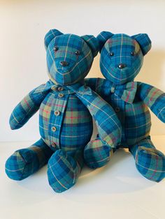 two blue teddy bears sitting next to each other on a white surface with one bear wearing a coat