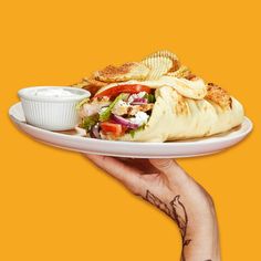 a hand holding a white plate with a taco and chips on it against an orange background