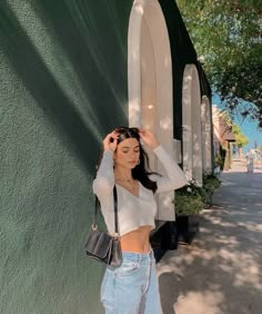 a woman standing in front of a green wall with her hand on her head and wearing high waist jeans