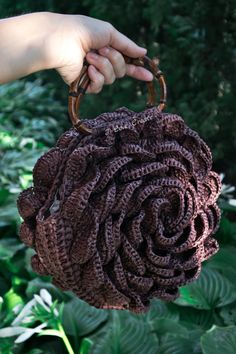 a hand holding a brown crocheted purse in front of green plants and trees