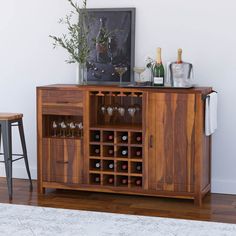 a wooden cabinet with wine glasses and bottles on it next to two stools in front of a white wall