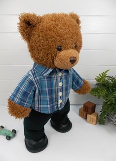 a brown teddy bear wearing a blue shirt and black pants standing next to a potted plant