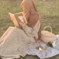 a woman in a hijab reading a book while sitting on the grass with a basket