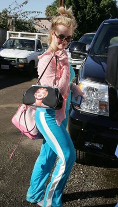 a woman in pink shirt and blue pants holding a purse while standing next to a car