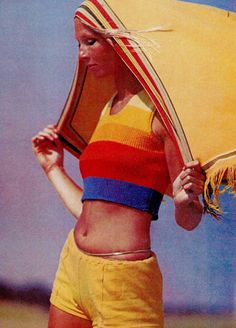 a woman in yellow shorts and a colorful top holding an umbrella over her head while standing on the beach