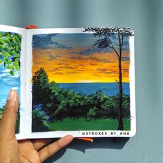 a hand holding an open book with a painting on the cover and trees in the background