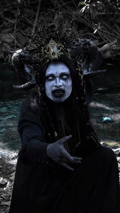 a woman with white makeup and black hair sitting on the ground next to a river