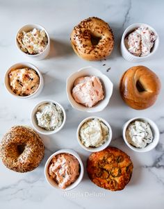 several different types of doughnuts in small bowls on a marble counter top with cream cheese and other toppings