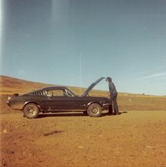 a man standing next to a black car in the middle of a dirt road with his hand on the hood