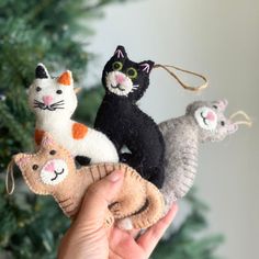 a hand holding three cat ornaments in front of a christmas tree