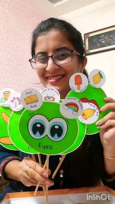 a woman holding up some green paper cut outs with eyes on them and stickers in front of her face