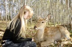 a woman sitting on the ground next to a deer in a forest filled with trees