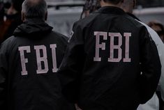 two men in black jackets with the letters fbi on them walking down a street