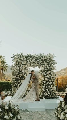 a bride and groom are kissing in front of an arch with white flowers on it