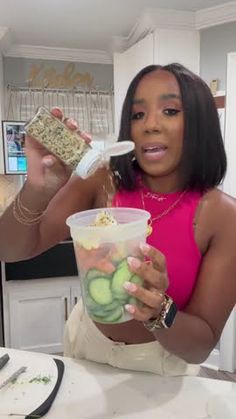 a woman holding up a plastic cup filled with cucumbers and popcorn while sitting at a table