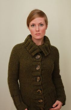 a woman is wearing a green sweater with buttons on the front and bottom, standing in front of a white wall