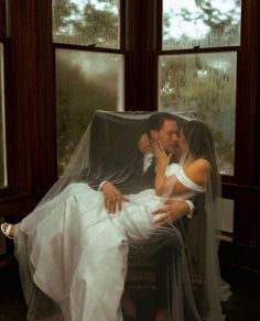 a bride and groom are sitting on a chair in front of two windows with rain coming through them