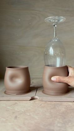 a hand is holding a brown vase on top of two small cups that are sitting next to each other