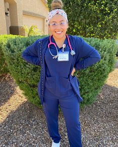 a woman wearing scrubs and a stethoscope standing in front of a bush