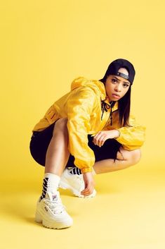 a woman in yellow jacket and white sneakers crouching on the ground with her legs crossed