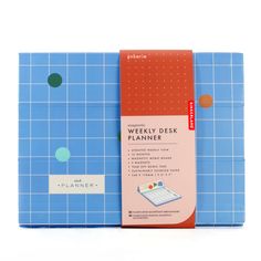 the weekly desk planner in blue and red is next to it's box on a white surface