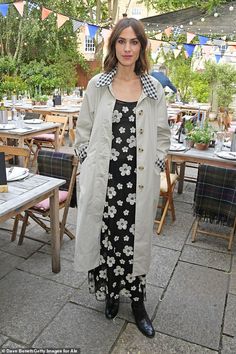 a woman standing in front of tables at an outdoor event wearing a trench coat and dress