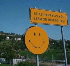 a yellow smiley face sign sitting on top of a metal pole next to a lush green field