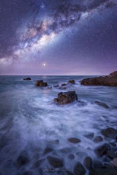 the night sky is filled with stars above the ocean and rocks in the foreground