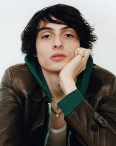 a young man wearing a brown leather jacket and green sweater with his hand on his chin