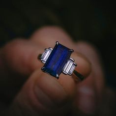 a person holding a ring with a blue stone in it's center and two baguetts on the side