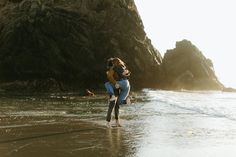 a woman carrying a child on her back walking along the beach near some rocks and water