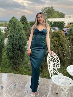 a woman in a green dress standing next to a white chair and table with trees behind her