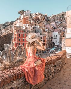 a woman in a dress and hat sitting on a stone wall looking at the city