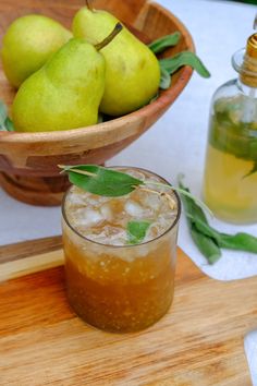 a wooden bowl filled with green pears next to a glass of iced tea on top of a cutting board